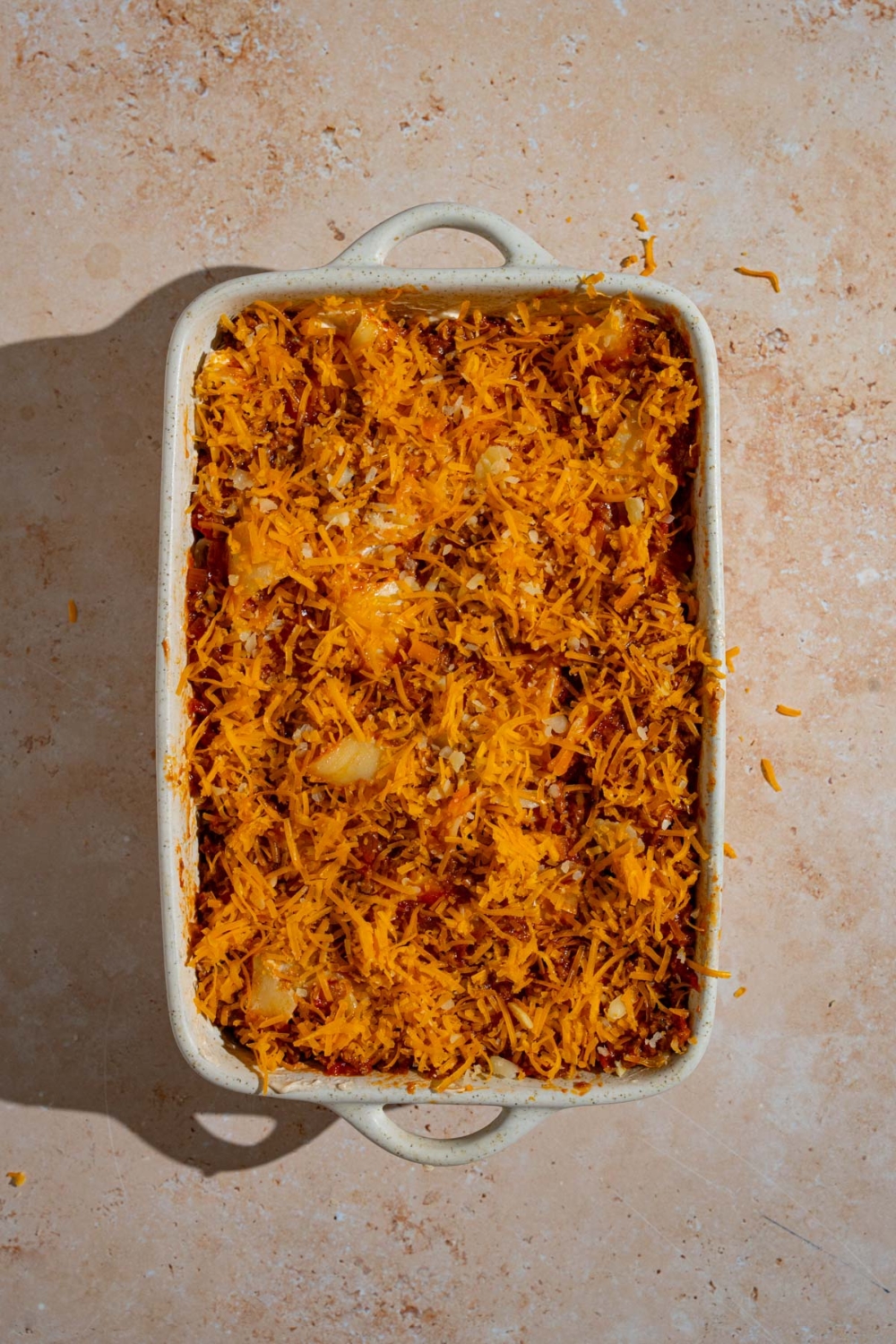 A baking dish with uncooked poor man's husband casserole topped with shredded colby jack cheese. The dish is on a tan counter.
