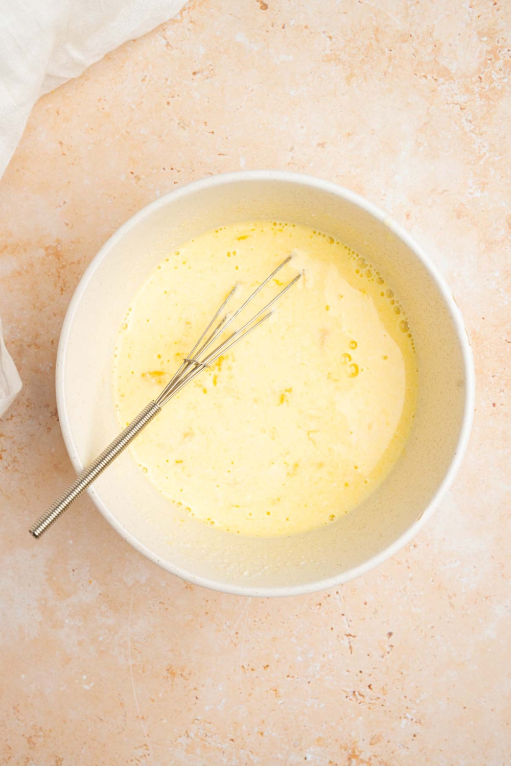A white bowl with a whisk combining ingredients to make fried cornbread. The bowl is on a tan counter with a white cloth napkin.