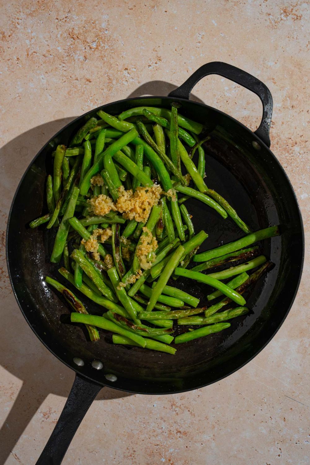A skillet with roasted green beans cooking with oil and garlic. The skillet is on a tan counter.