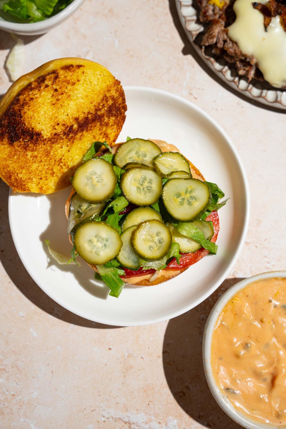 An open In-N-Out burger with the bottom bun layered with sauce, sliced tomato, shredded lettuce, and sliced pickles. The plate is on a tan counter with a bowl of burger sauce and plate of burger patties.