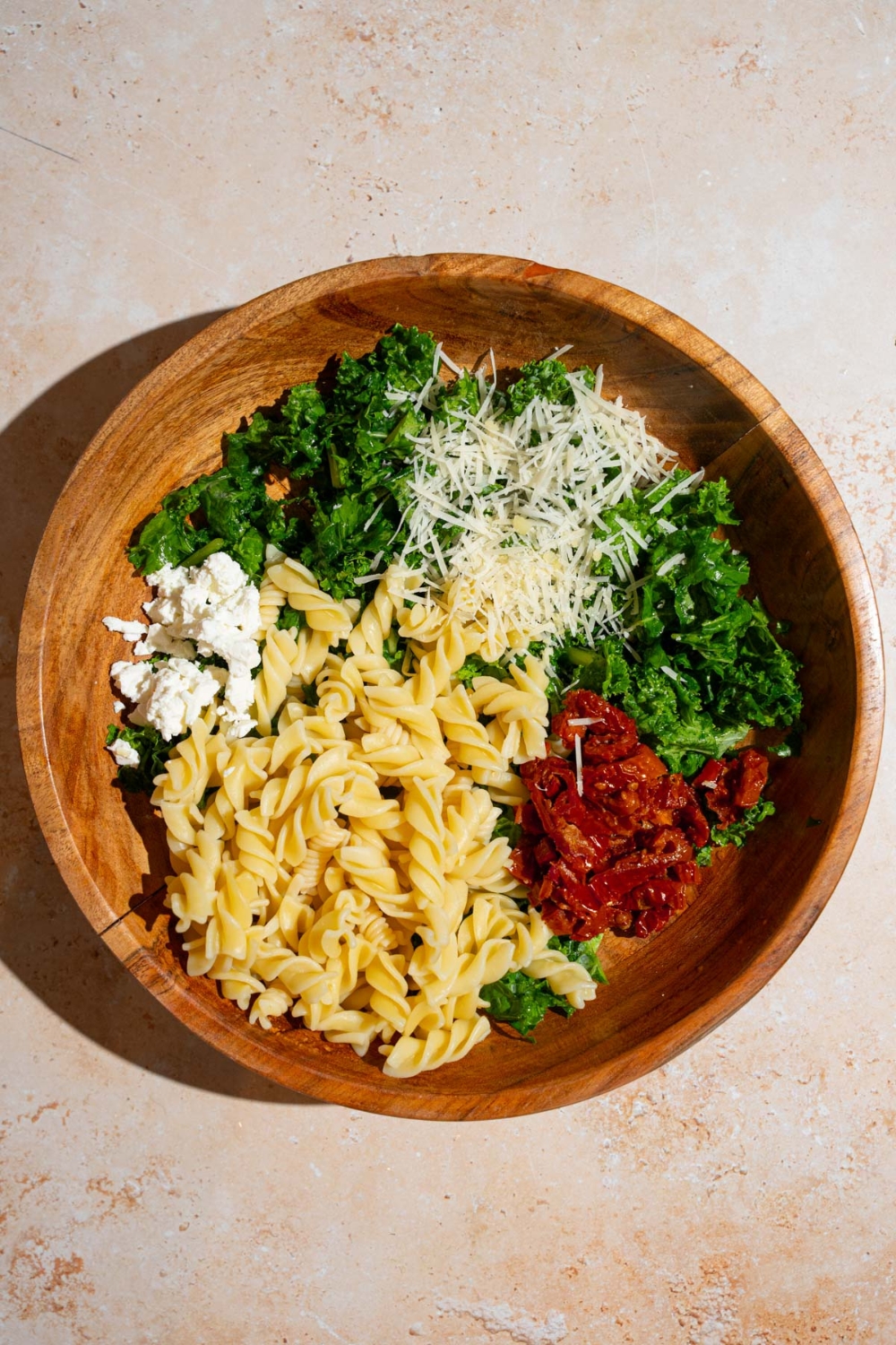 A wooden bowl with ingredients to make Mediterranean pasta salad including kale, rotini pasta, sundried tomatoes, feta cheese, and grated parmesan. The bowl is on a tan counter.