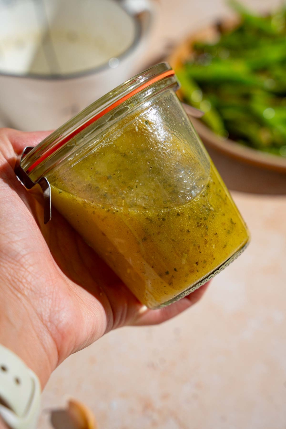A hand holding a sealed jar of copycat Texas Roadhouse italian dressing. There is a tan counter with a plate of green beans blurred in the background.