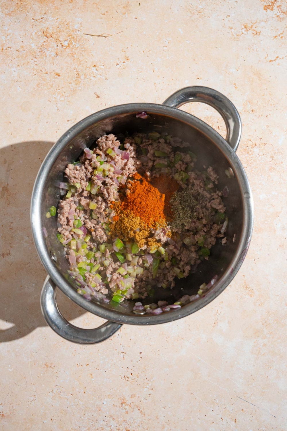 A stock pot with browned ground beef and sausage with sautéd green peppers, celery, and onion. The pot is on tan counter.