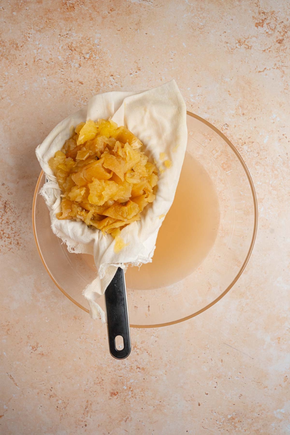 A glass bowl with strained apple juice. There is a mesh strainer lined with cloth filled with cooked apple pieces. The bowl is on a tan counter.