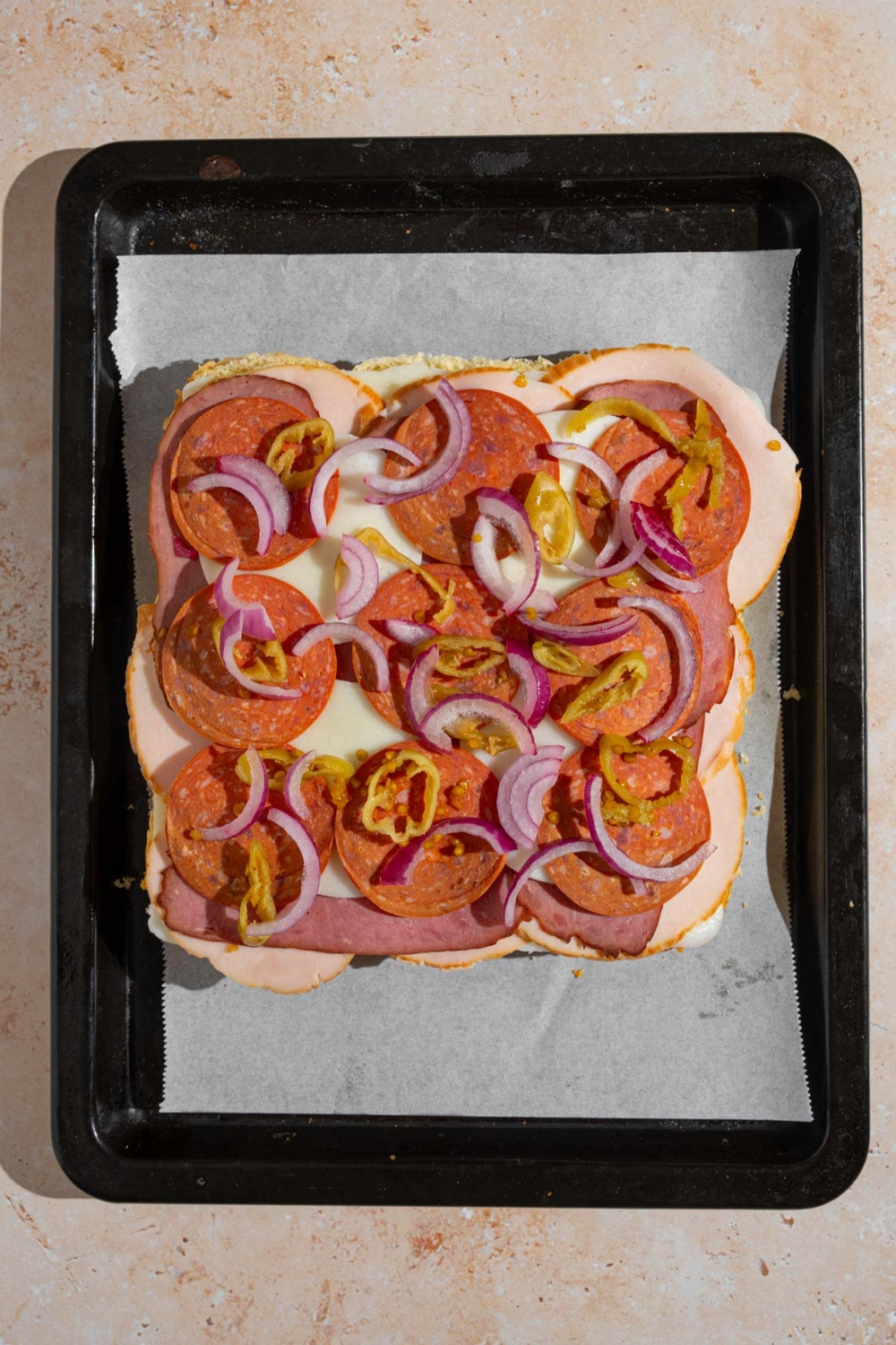 Nine open Italian sliders topped with sliced turkey, salami, cheese, pepperocinis, and sliced red onion. The sliders are on a baking sheet lined with parchment paper.