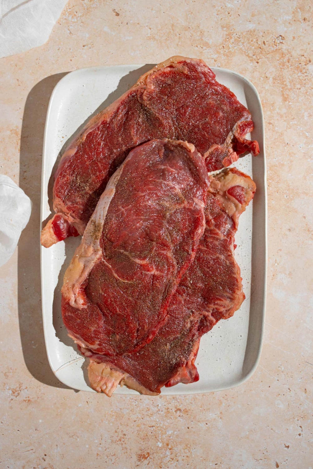 Three tenderized uncooked sirloin steaks on a white platter. The platter is on a tan counter.