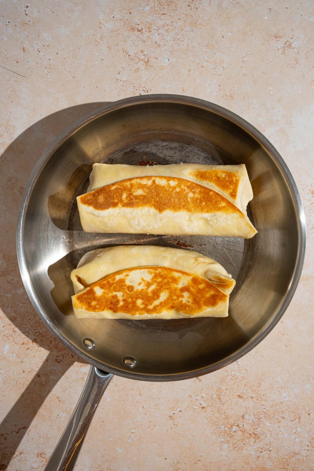 A skillet with two cheesy garlic chicken wraps cooking in oil. The skillet is on a tan counter.
