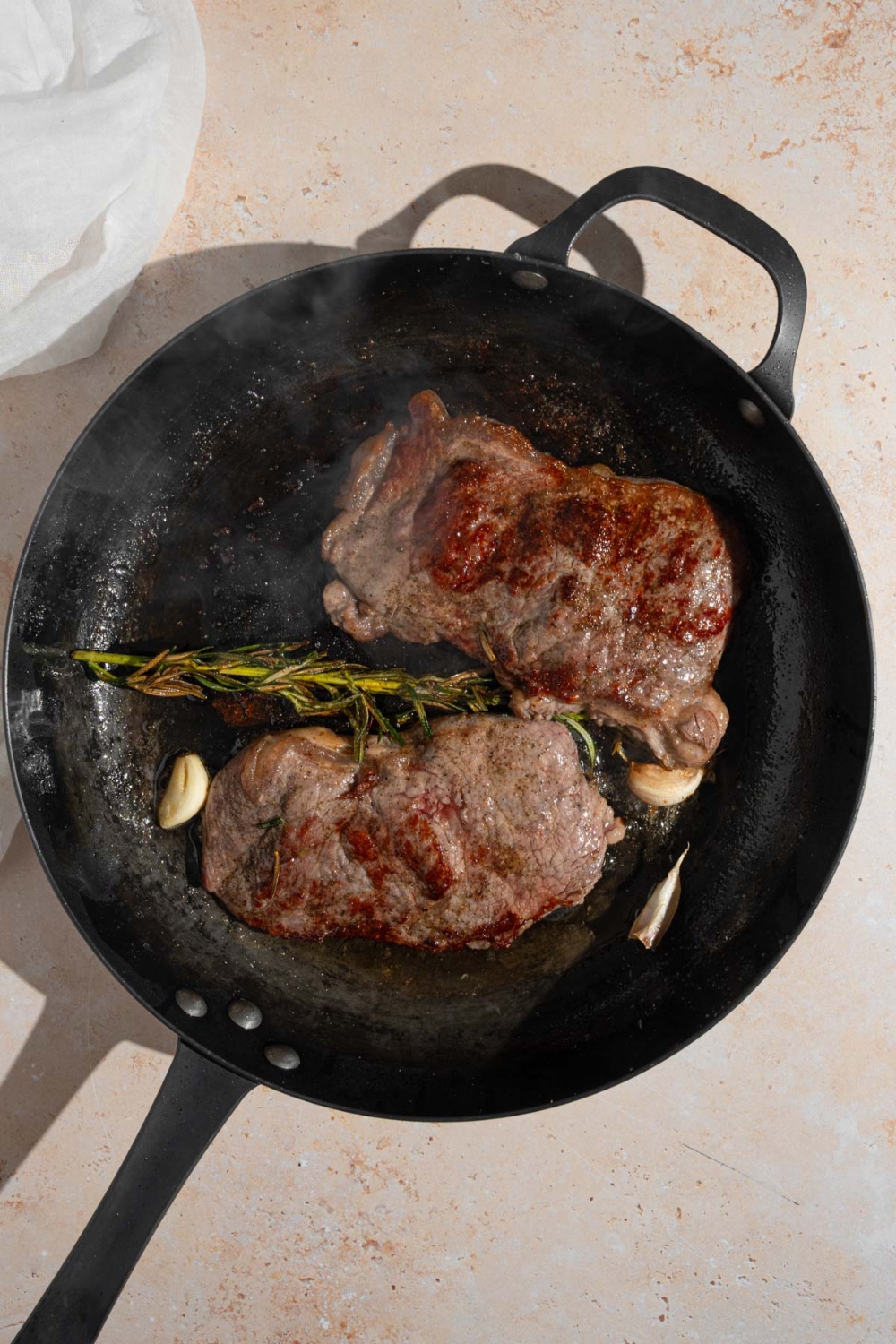 A cast iron skillet with two cooked sirloin steaks with a sprig of rosemary and garlic. The pan is on a tan counter with a white cloth napkin.
