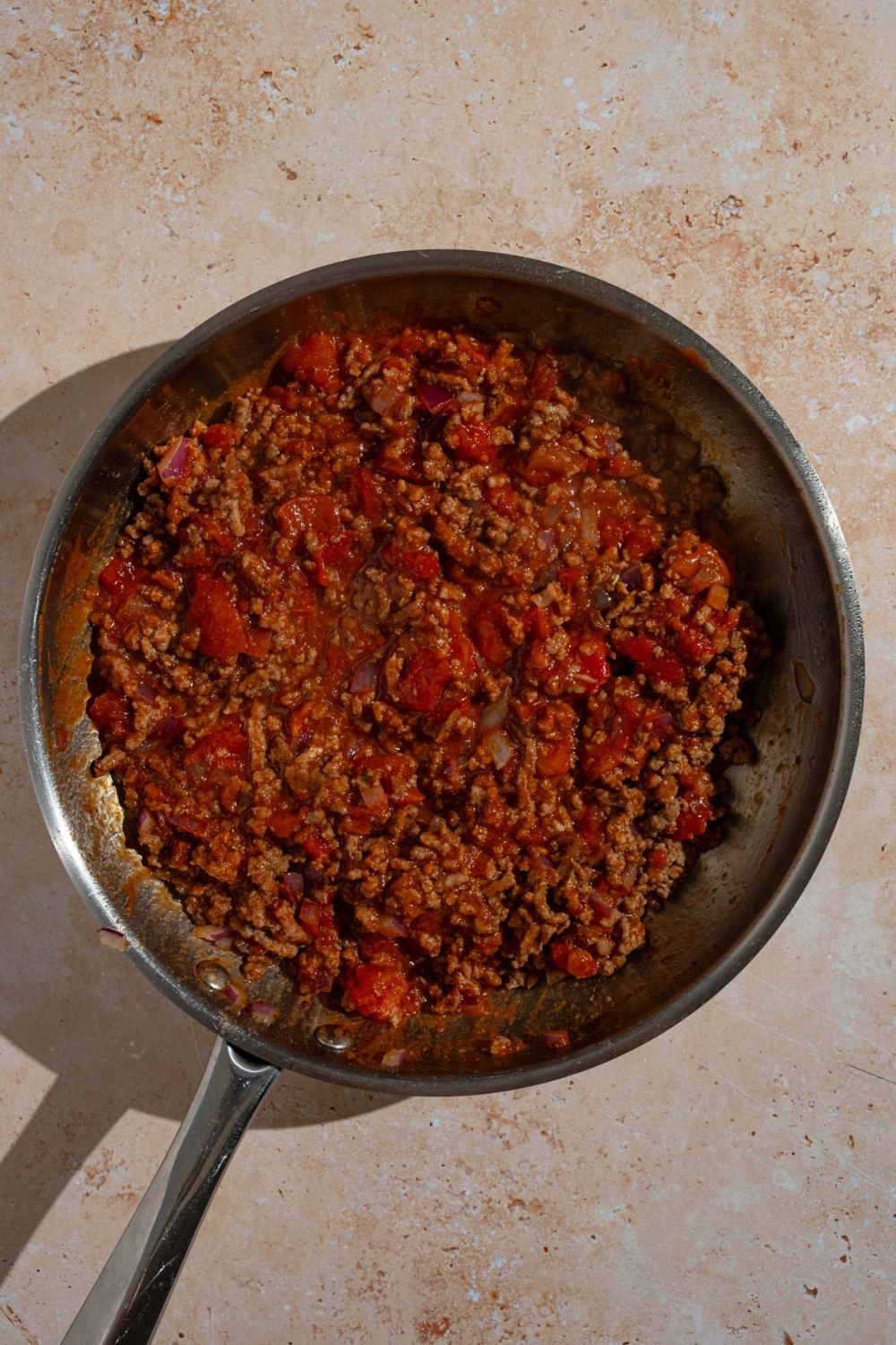 A skillet with seasoned browned beef mixed with tomato sauce. The skillet is on a tan counter.