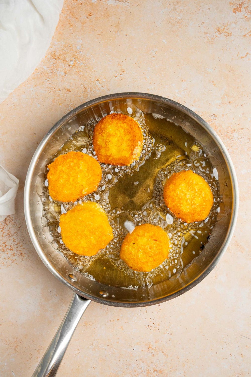 A skillet with several pieces of cornbread frying in oil. The skillet is on a tan counter with a white cloth napkin.