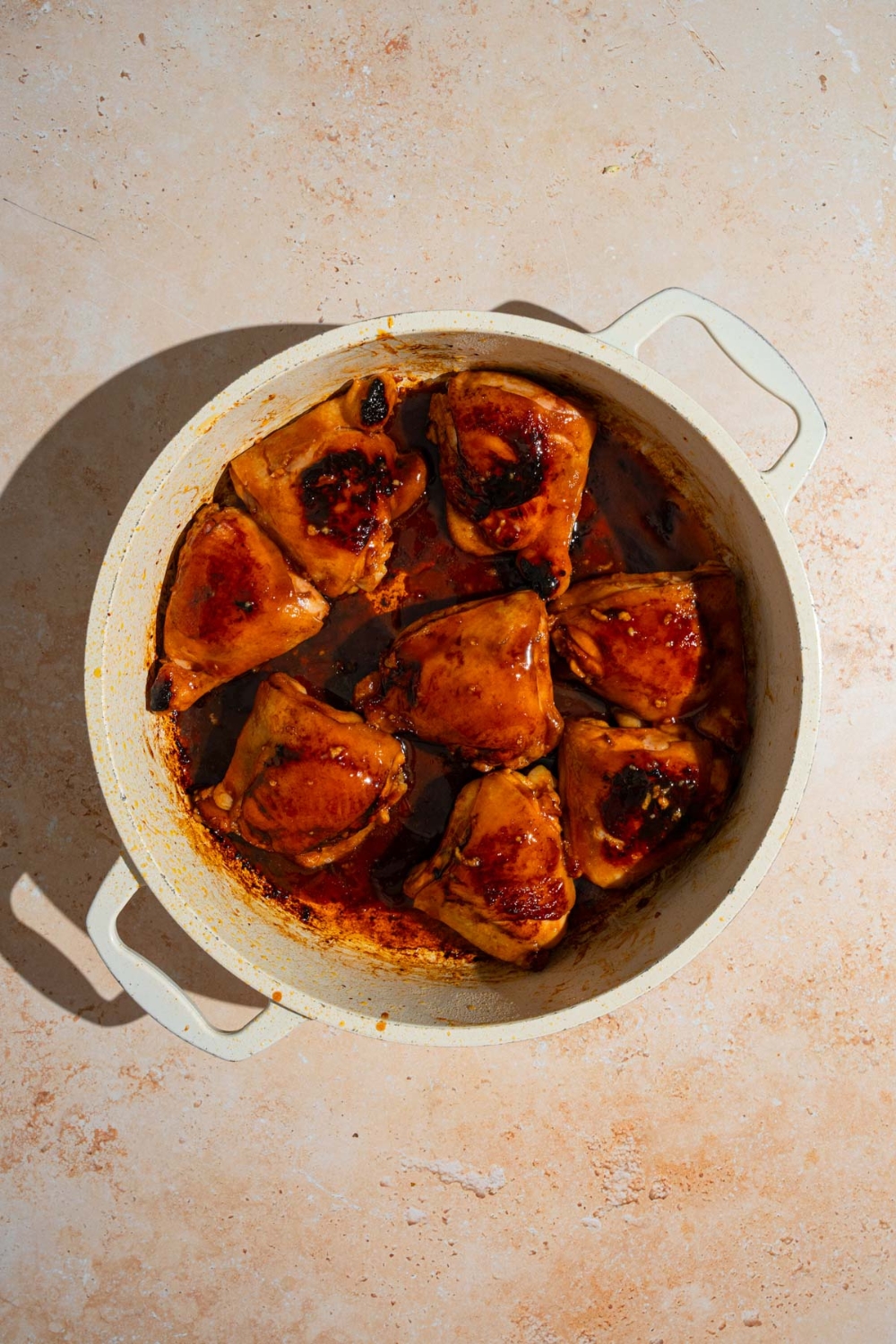 A large ceramic pot with seared chicken thighs covered with marinade. The pot is on a tan counter.