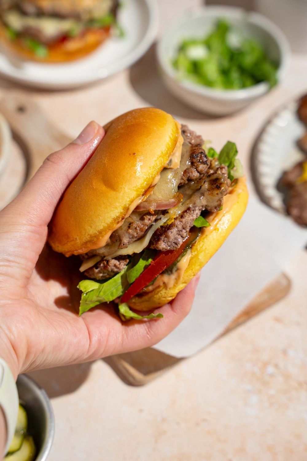 A hand holding a copycat In-N-Out burger. There is a board lined with parchment paper and bowl of lettuce blurred in the background.