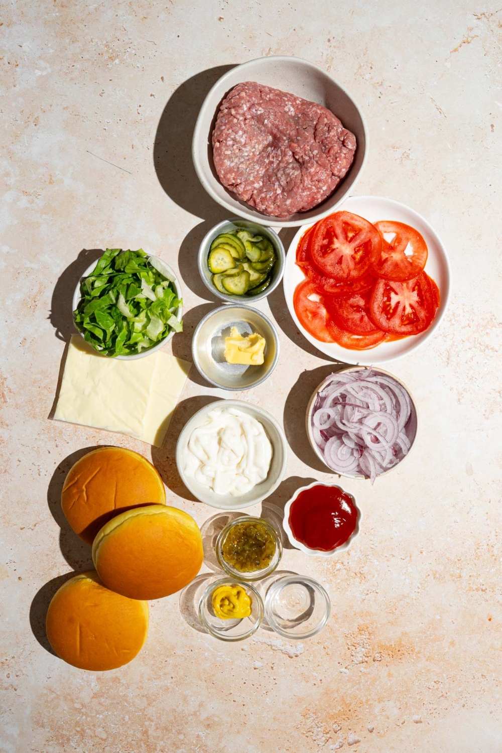 An overhead shot of several bowls in various sizes containing ingredients to make copycat In-N-Out burgers including burger buns, ground beef, sliced tomato, sliced red onion, shredded lettuce, American cheese, sliced pickles, mayo, ketchup, mustard, mayo, and seasonings.