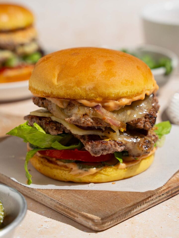 A copycat In-N-Out burger on a wooden board lined with parchment paper. The board is on a tan counter with an additional burger on a plate and small bowl of lettuce.