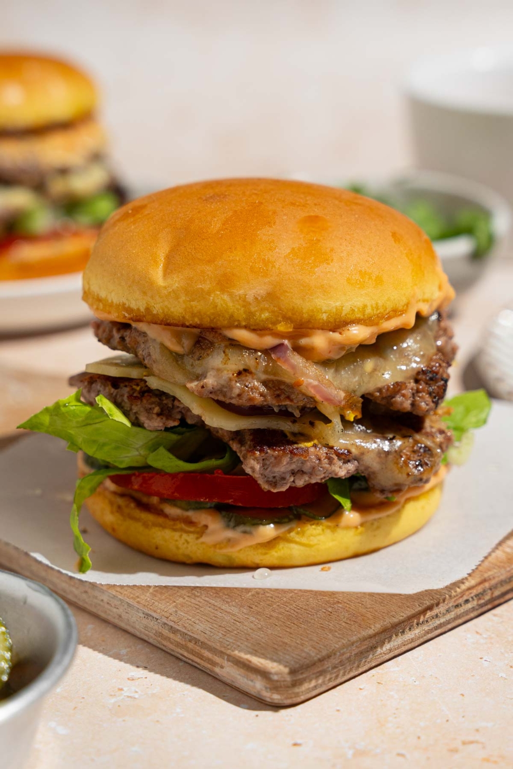 A copycat In-N-Out burger on a wooden board lined with parchment paper. The board is on a tan counter with an additional burger on a plate and small bowl of lettuce.