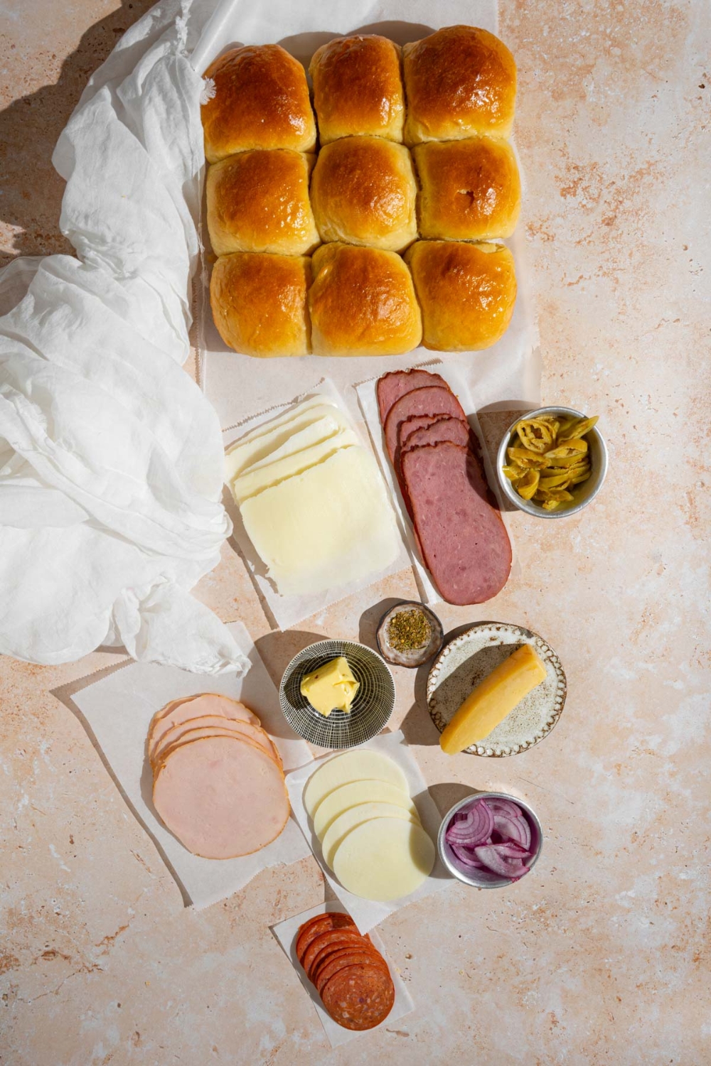 An overhead shot of ingredients to make Italian sliders including slider rolls, sliced turkey, sliced ham, sliced pepperoni, sliced cheese, sliced onion, and sliced pepperoncini.