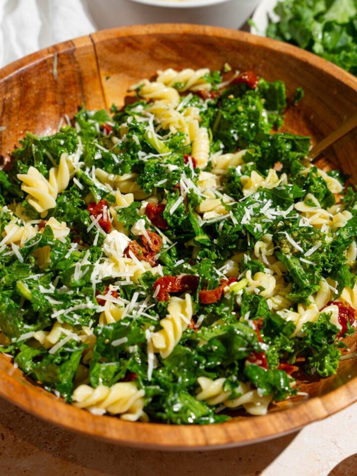 Mediterranean pasta salad served in a wooden bowl garnished with grated parmesan. The salad includes kale, rotini pasta, and sundried tomatoes. The bowl is on a tan counter with a plate of chopped kale and bowl of pasta.