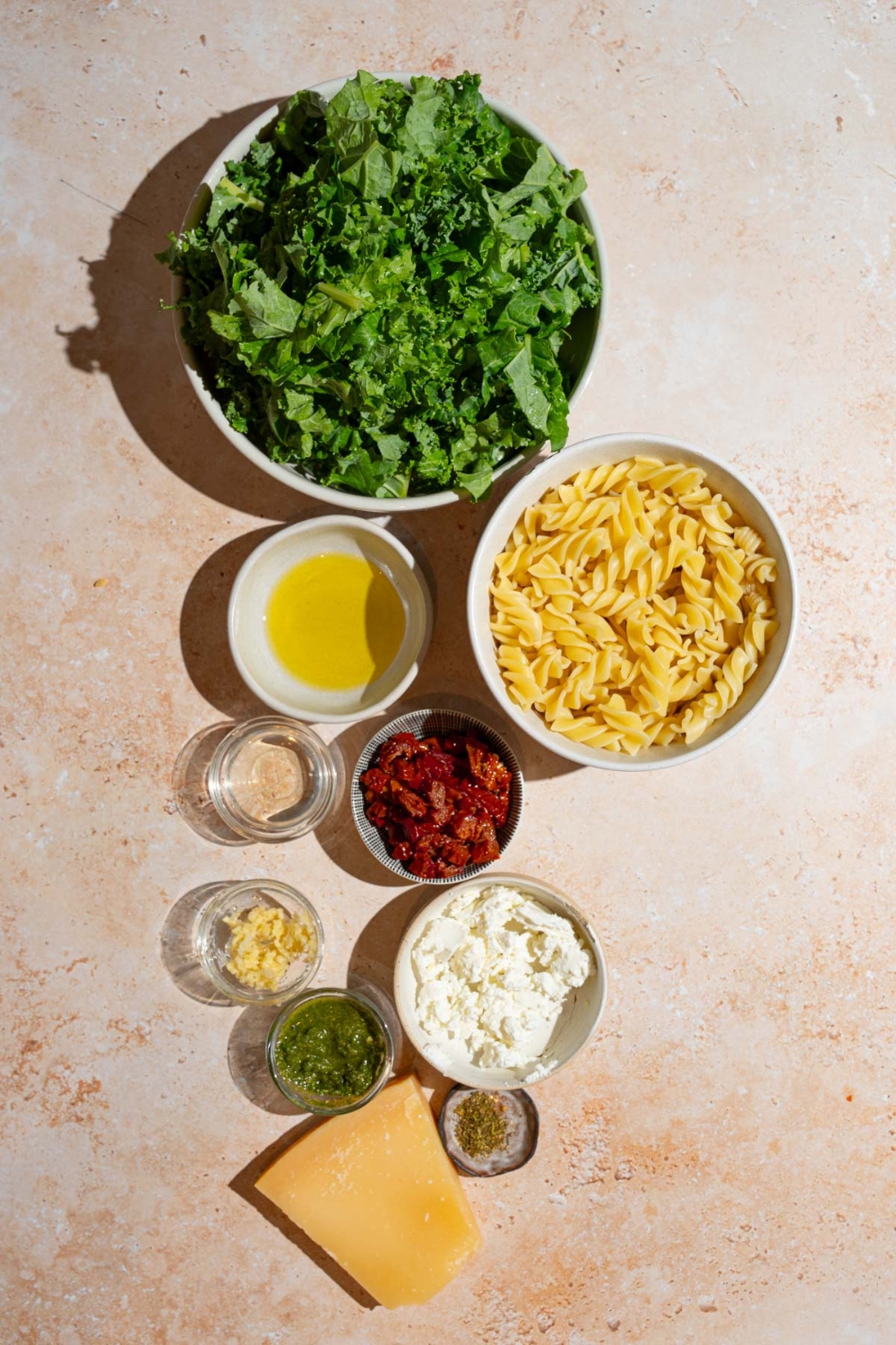 An overhead shot of several bowls in various sizes containing ingredients to make Mediterranean pasta salad including chopped kale, rotini pasta, sundried tomatoes, olive oil, pesto, balsamic vinegar, garlic, feta cheese, and seasonings.