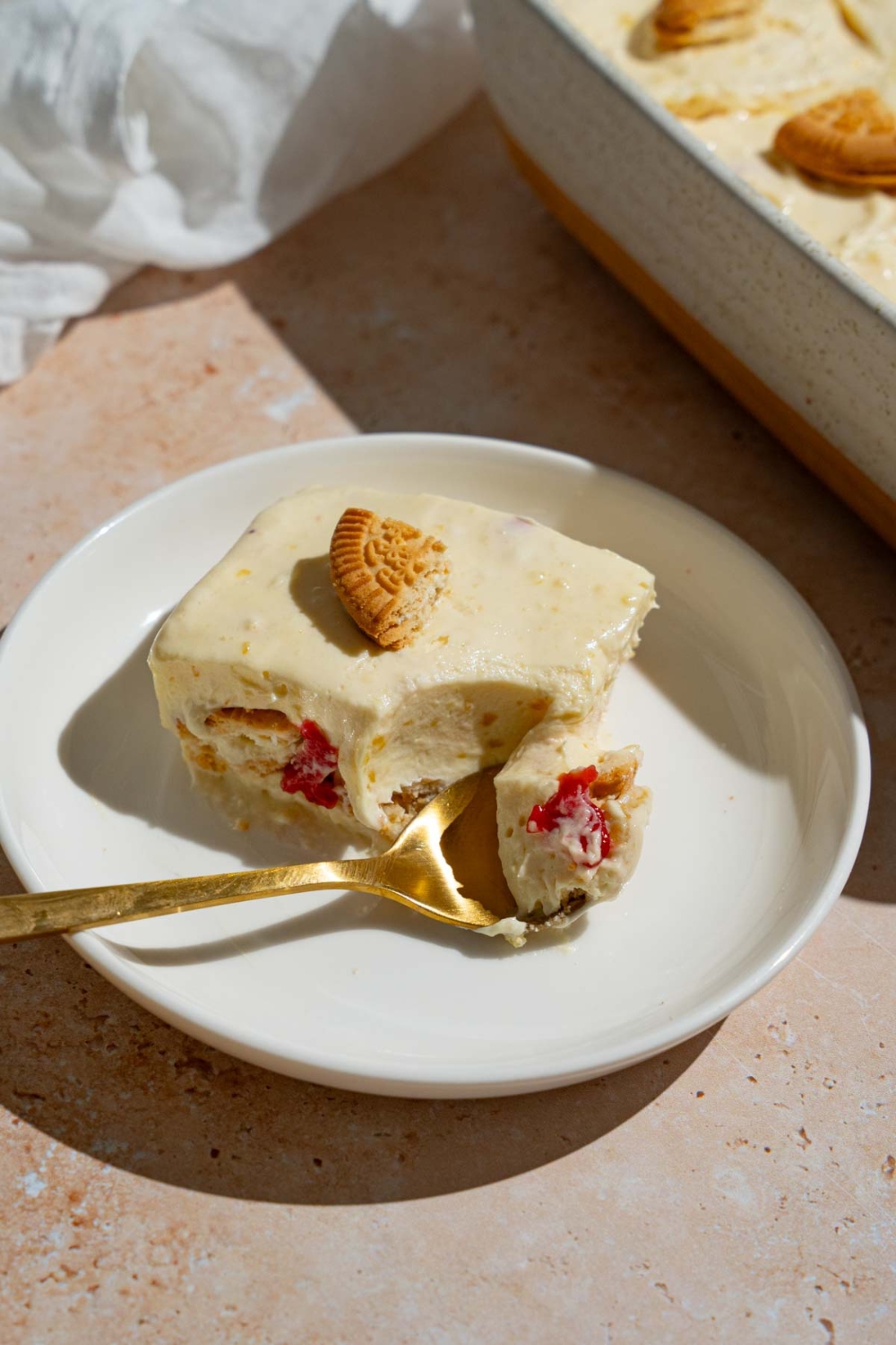 A slice of strawberry banana puding topped with a piece of a vanilla cookie. The pudding is on a white plate with a spoon taking a bite of pudding. The plate is on a tan counter with a baking dish with pudding.