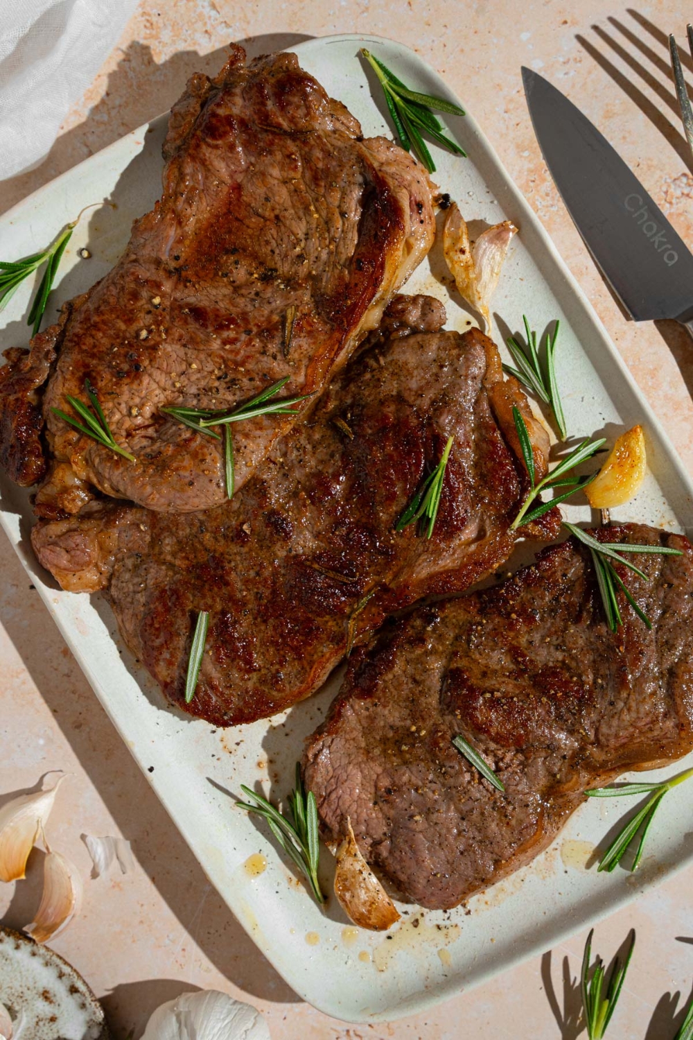 A tray of pan-fried seasoned sirloin steaks. The steaks are on a white platter with fresh rosemary and garlic. The tray is on a tan counter with a knife.