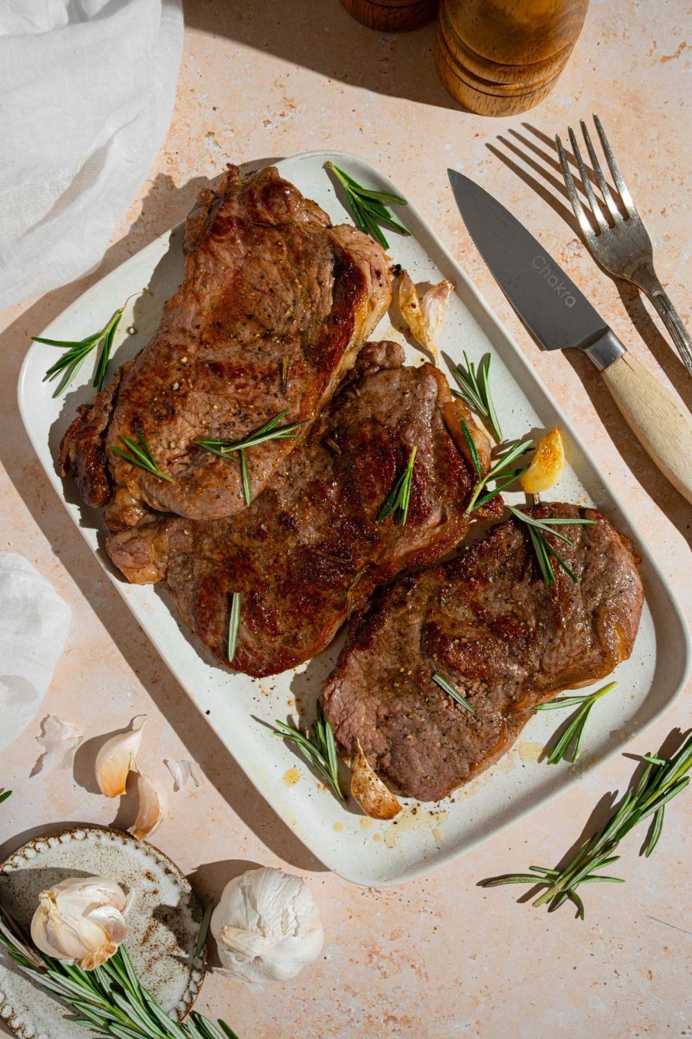 A tray of pan-fried seasoned sirloin steaks. The steaks are on a white platter with fresh rosemary and garlic. The tray is on a tan counter with a fork, knife, and small plate of garlic.