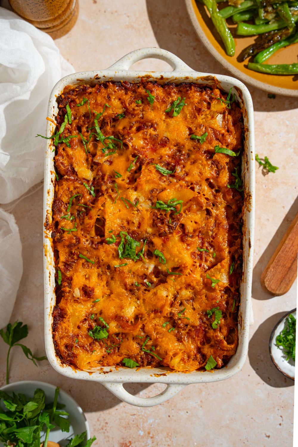 A baking dish with poor man's husband casserole garnished with fresh parsley. The dish is on a tan counter with a plate of garnishes and white cloth napkin.
