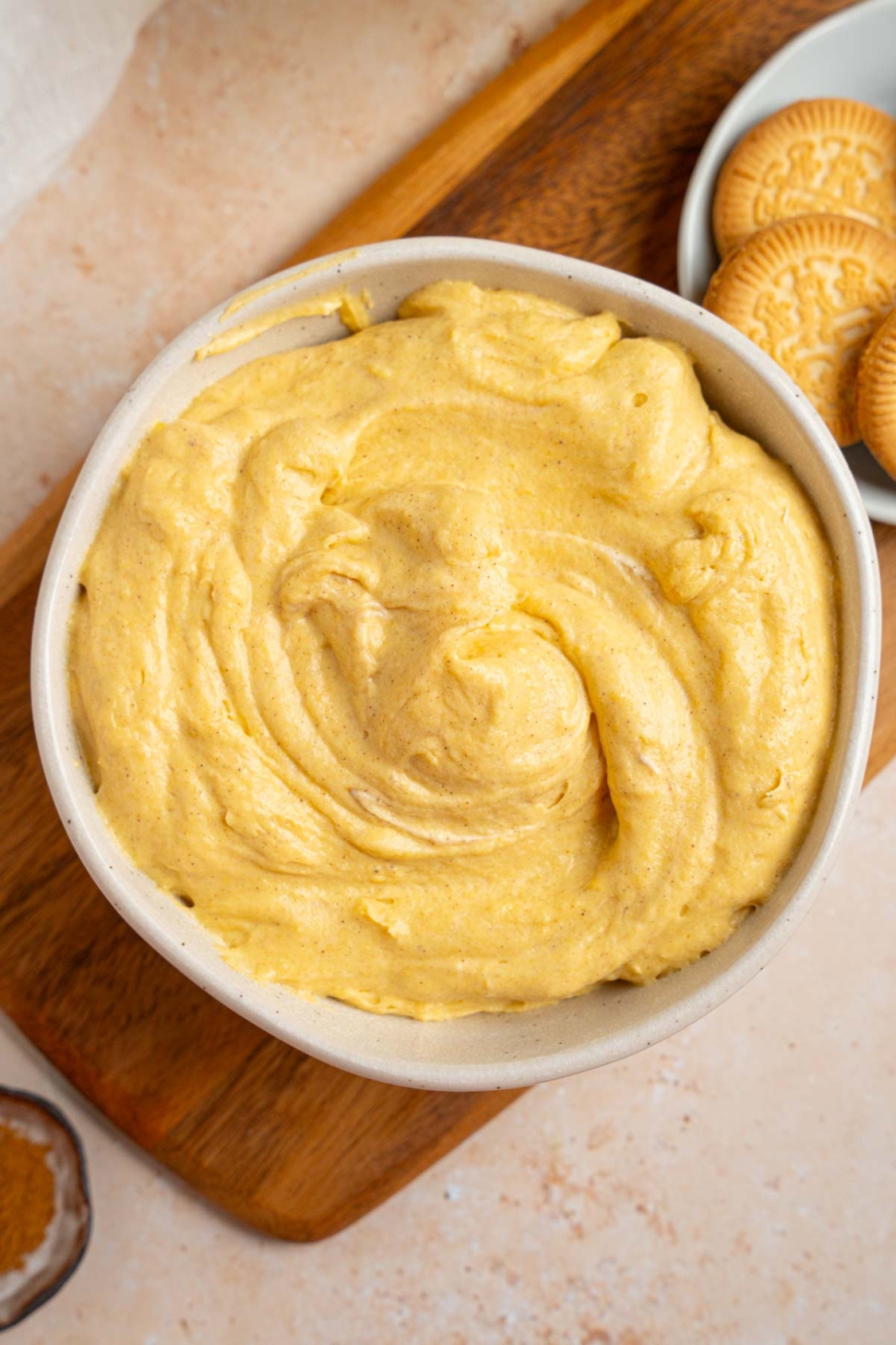 A white bowl with pumpkin fluff. The bowl is on a wooden board with a small bowl of vanilla cookies. The board is on a tan counter with a small ramekin of pumpkin spice.
