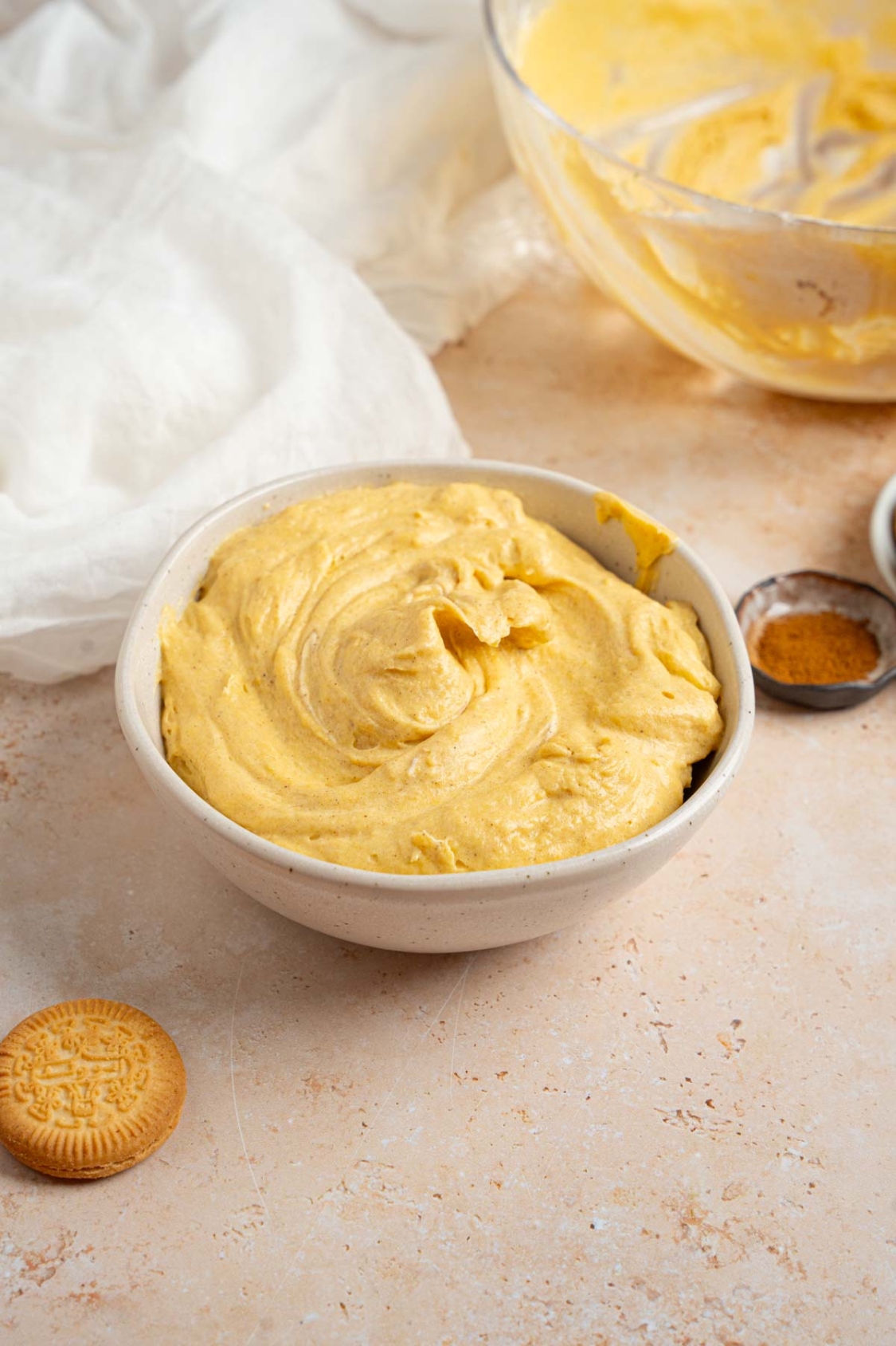 A white bowl with pumpkin fluff on a tan counter with a vanilla cookie, ramekin of pumpkin spice, and glass bowl of fluff.
