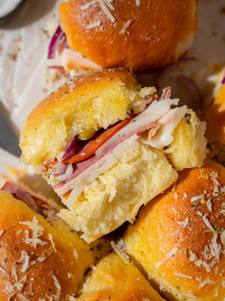 Italian sliders topped with grated cheese and seasonings served on pieces of parchment paper. One of the sliders is on its side.