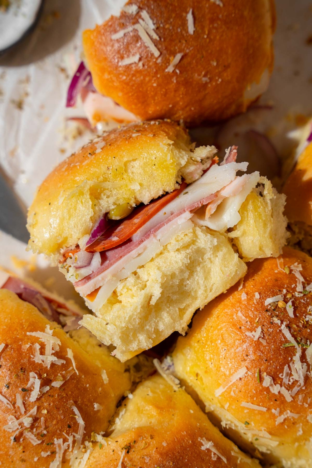 Italian sliders topped with grated cheese and seasonings served on pieces of parchment paper. One of the sliders is on its side.