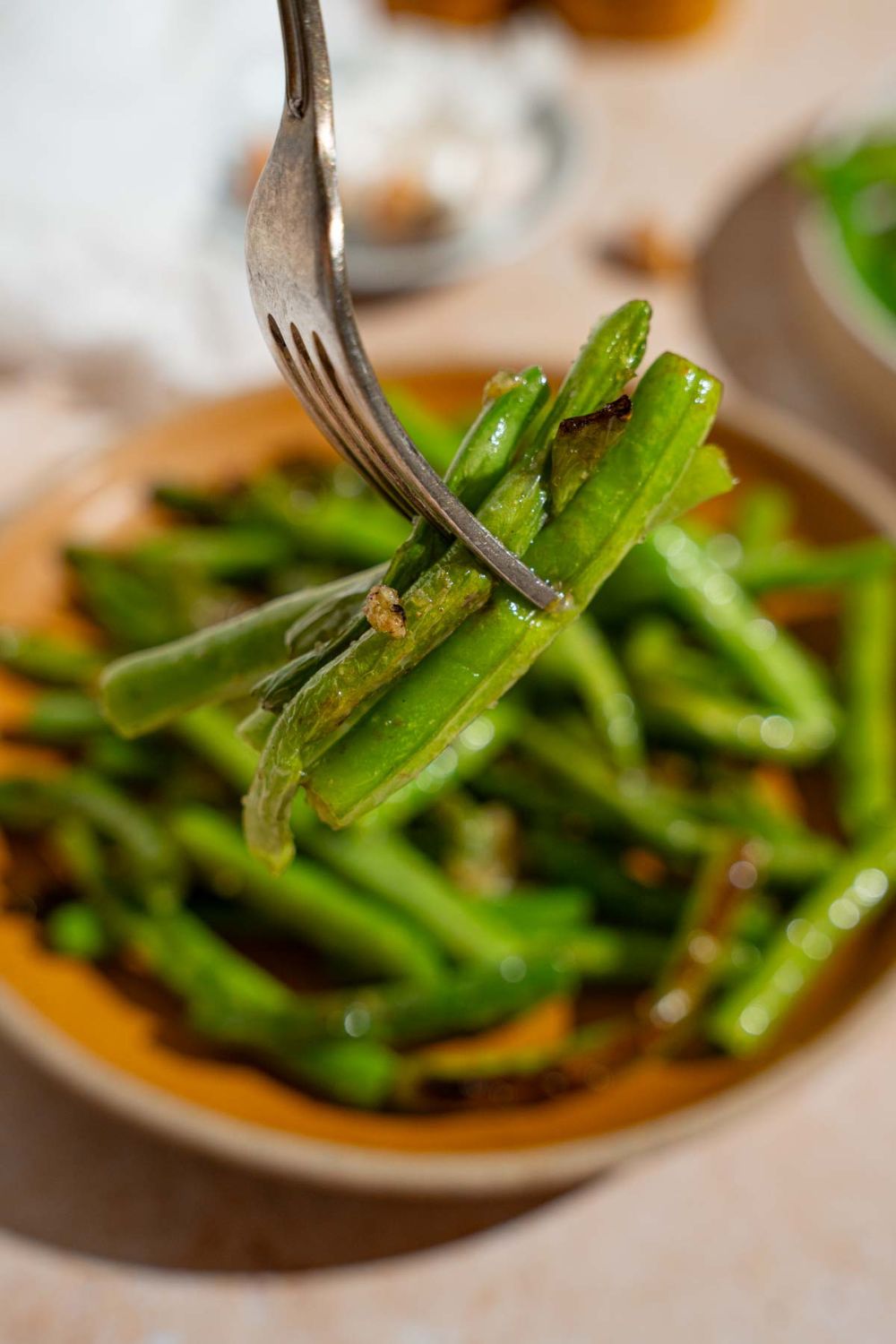 A close up of a fork with a bite of copycat Din Tai Fung green beans. There is an orange plate with green beans blurred in the background.