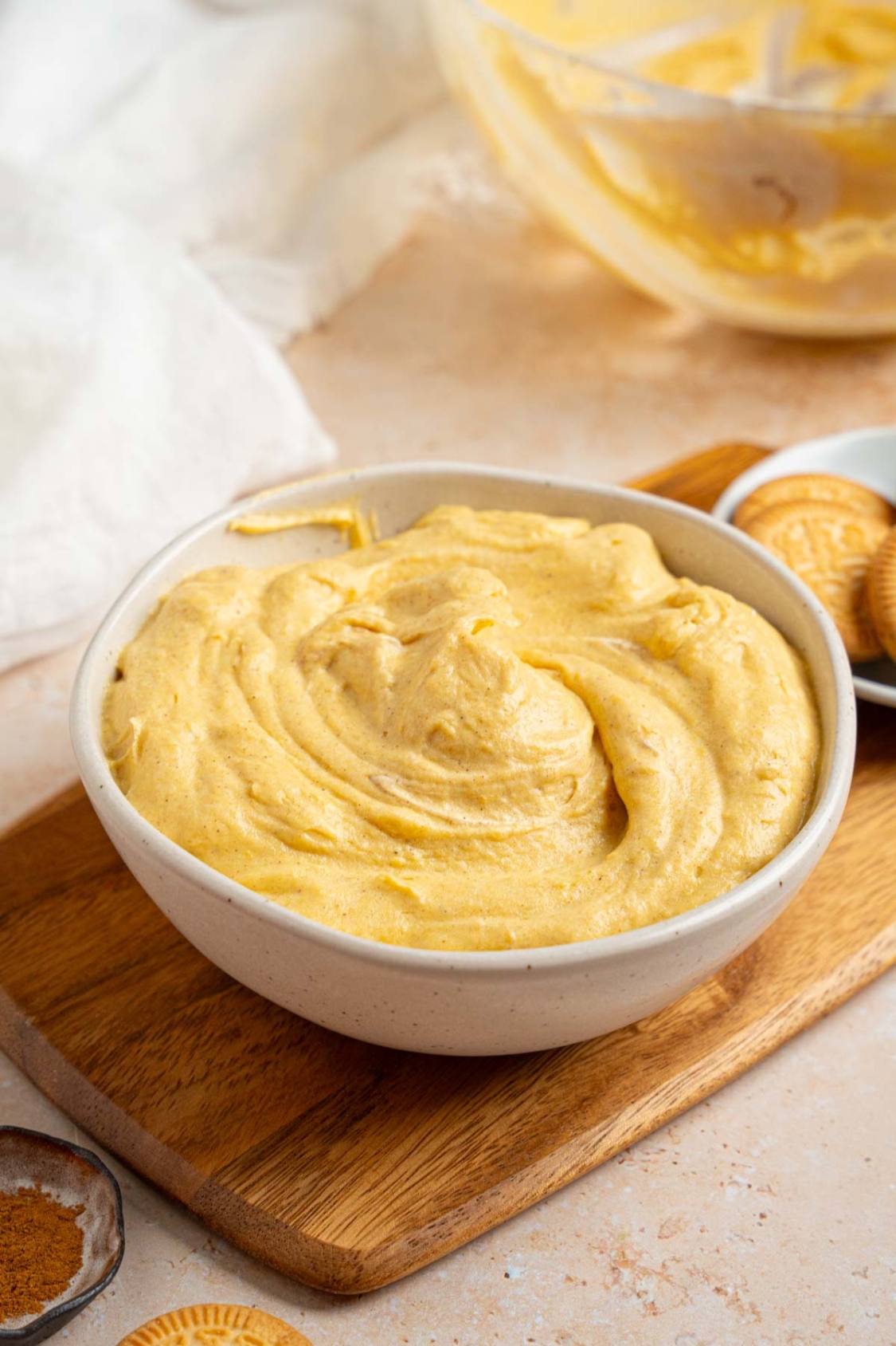A white bowl with pumpkin fluff. The bowl is on a wooden board with a small bowl of vanilla cookies. The board is on a tan counter with a small ramekin of pumpkin spice and glass bowl of fluff.