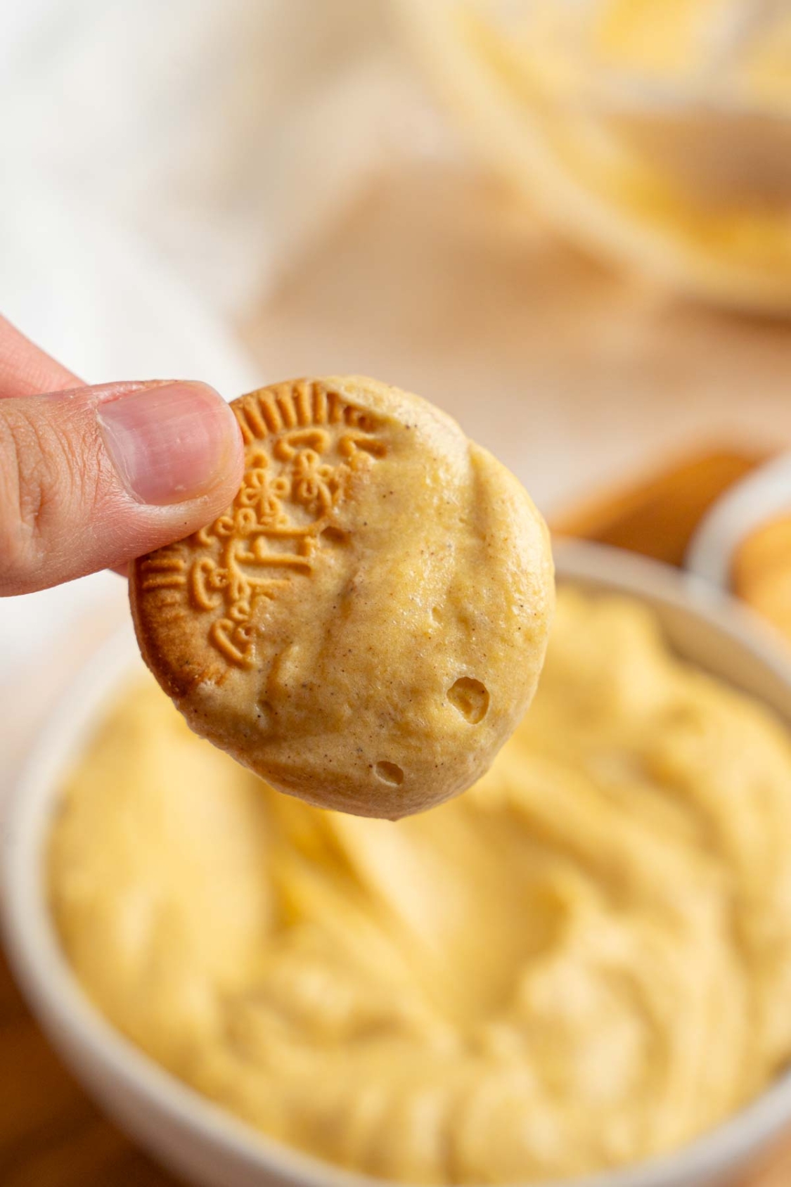 A close up of a hand holding a vanilla cookie dipped in pumpkin fluff. There is a bowl of fluff on a wooden board with a white cloth napkin blurred in the background.