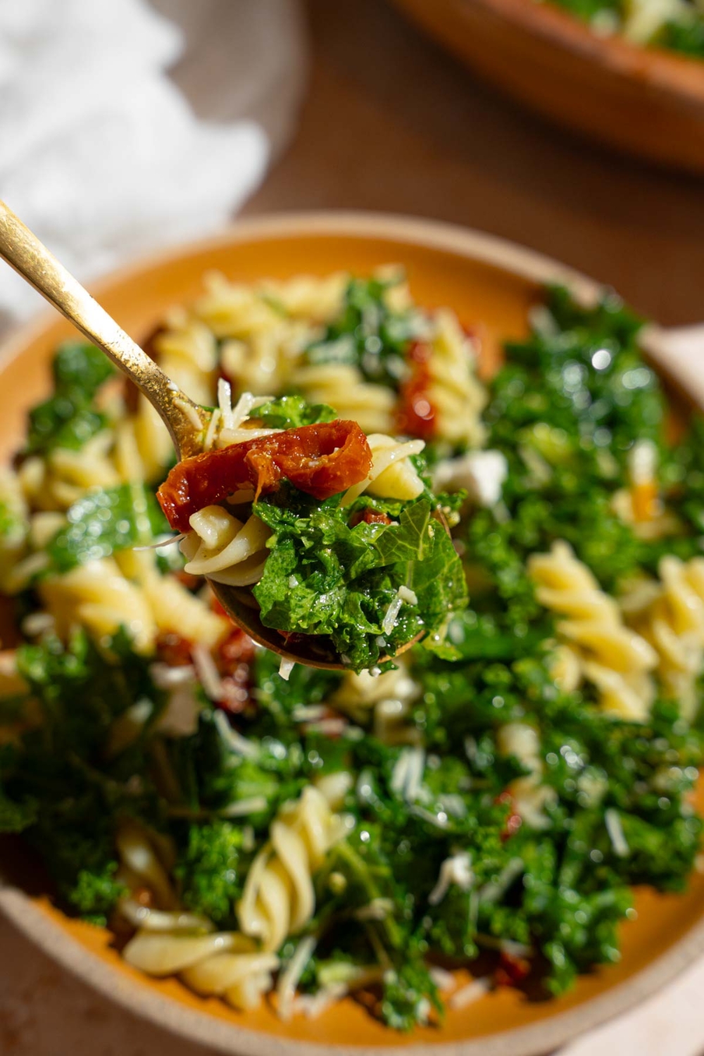 A close up of a fork with a bite of Mediterranean pasta salad. There is a wooden bowl of pasta salad on a tan counter with a white cloth napkin blurred in the background.