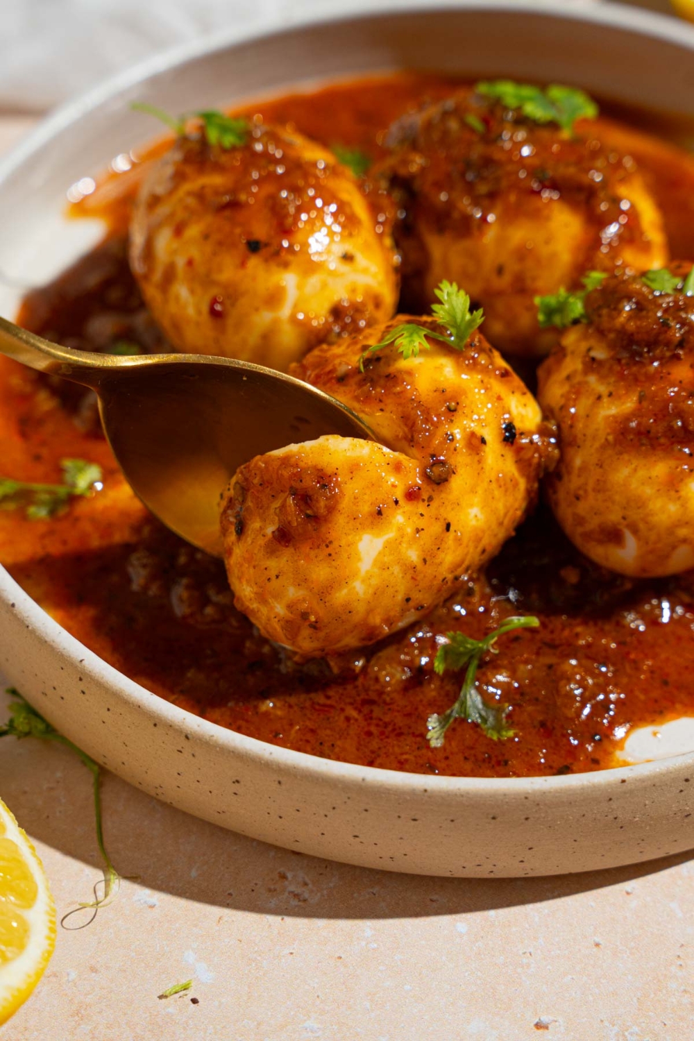 A white plate with four cajun boiled eggs covered in cajun sauce garnished with fresh parsley. A spoon is taking a bite from one of the eggs.
