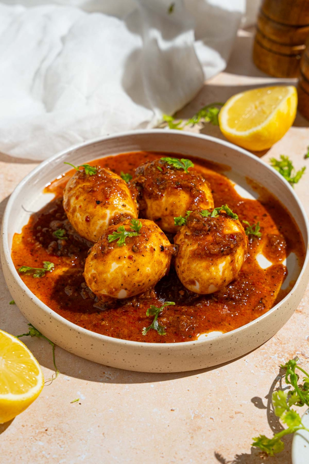 A white plate with four cajun boiled eggs covered in cajun sauce garnished with fresh parsley. The plate is on a tan counter with a sliced lemon, white cloth napkin, and plate of garnishes.