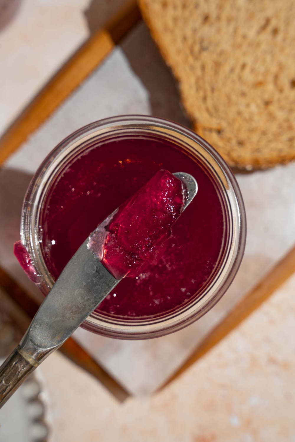 A knife in a jar of chokecherry jelly. The jar is on a wooden board lined with parchment paper with a slice of toast.