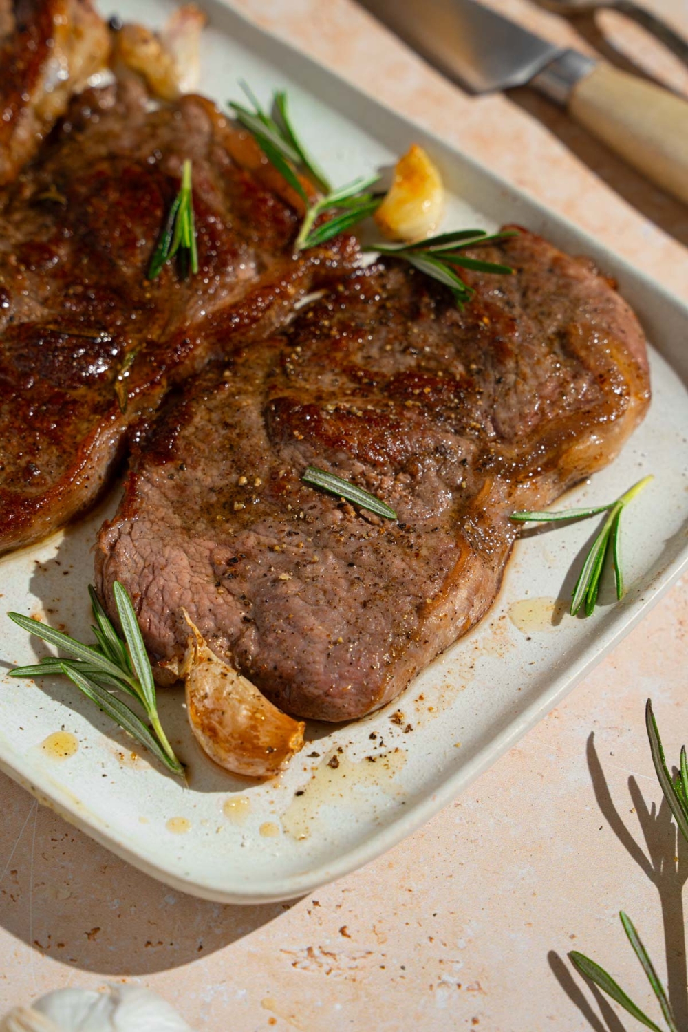 A tray of pan-fried seasoned sirloin steaks. The steaks are on a white platter with fresh rosemary and garlic. The tray is on a tan counter with a knife.