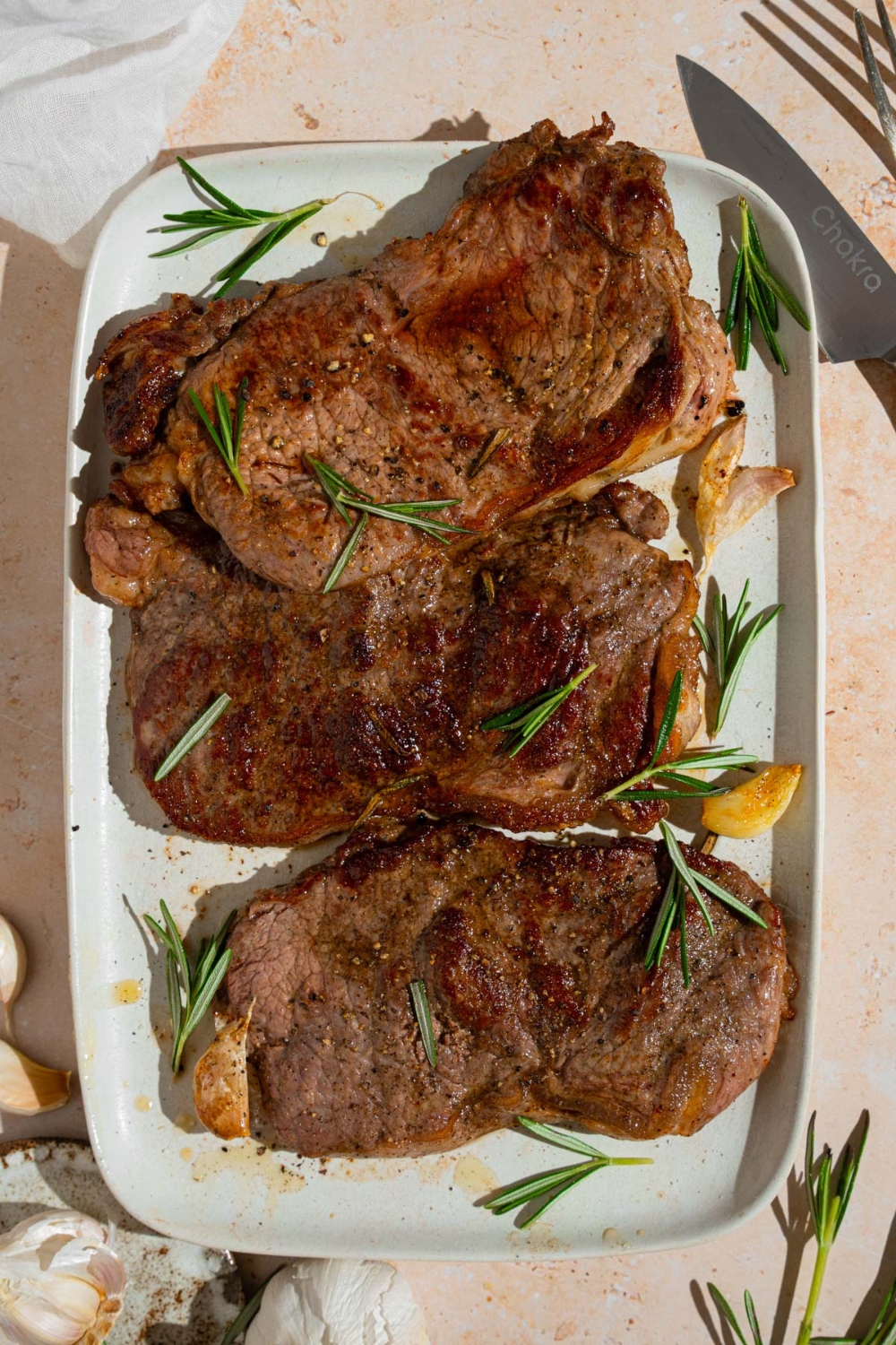 A tray of pan-fried seasoned sirloin steaks. The steaks are on a white platter with fresh rosemary and garlic. The tray is on a tan counter with a knife and small plate of garlic.