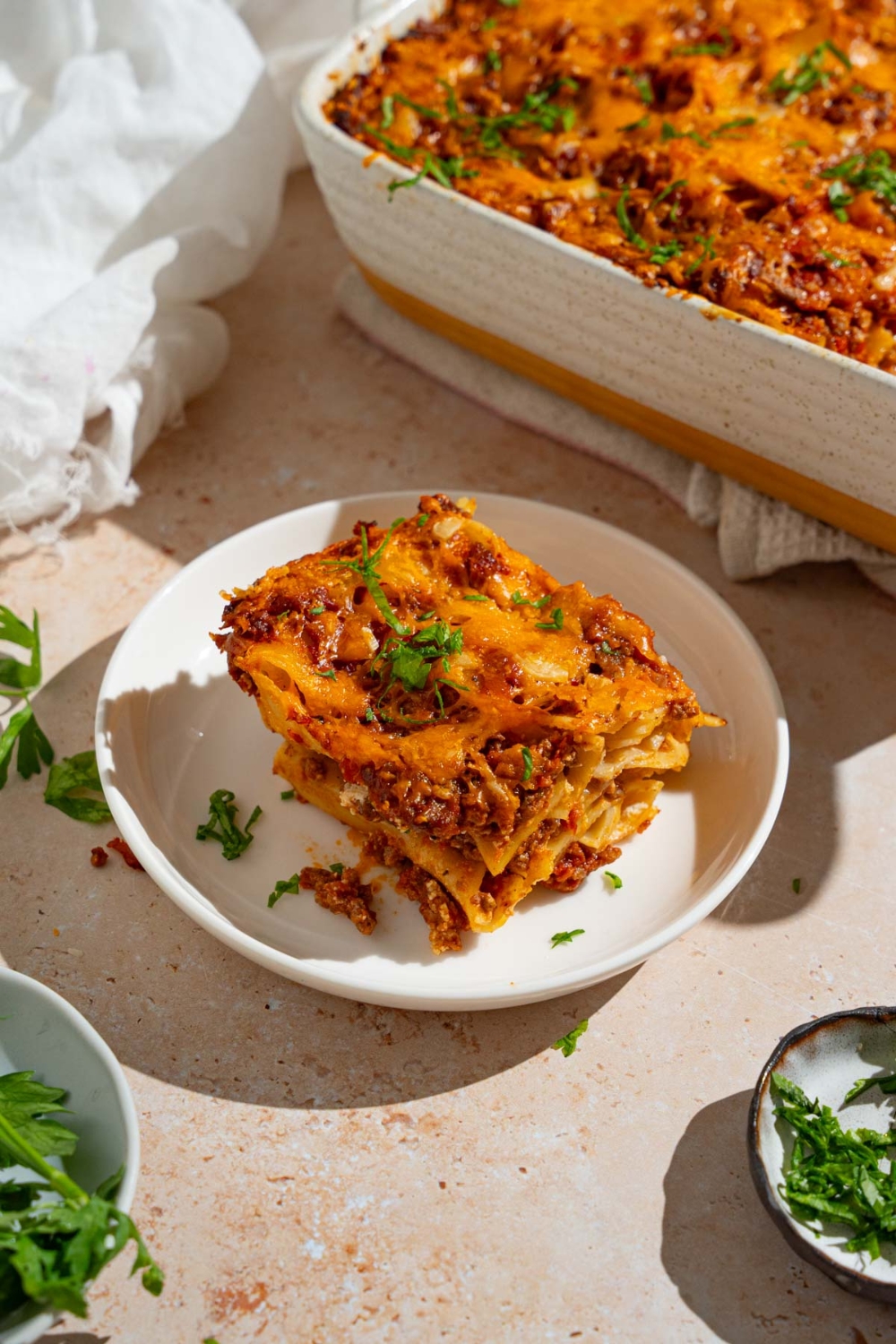 A slice of poor man's husband casserole garnished on a white plate garnished with fresh parsley. The plate is on a tan counter with a tray of casserole and small plates with garnishes.