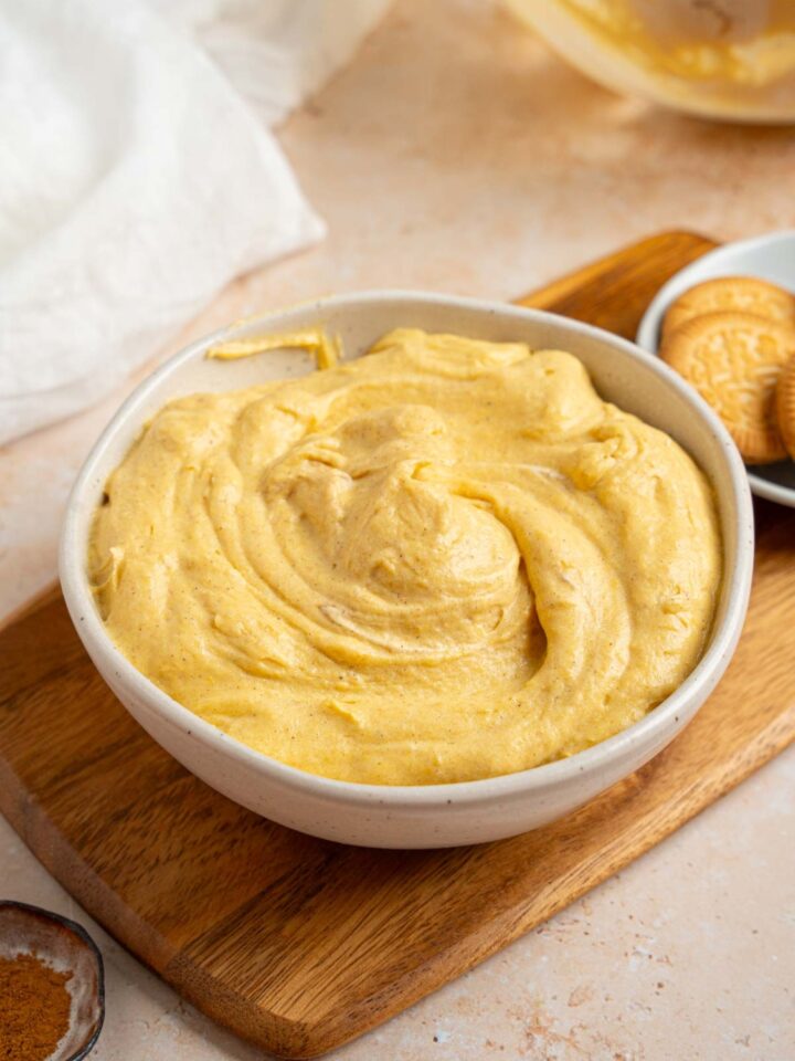A white bowl with pumpkin fluff. The bowl is on a wooden board with a small bowl of vanilla cookies. The board is on a tan counter with a small ramekin of pumpkin spice and glass bowl of fluff.