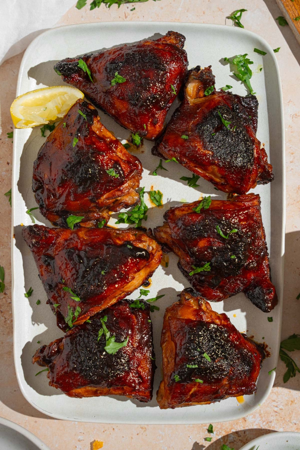 Several sticky chicken thighs served on a white platter garnished with fresh parsley and lemon. The platter is on a tan counter with a white cloth napkin.