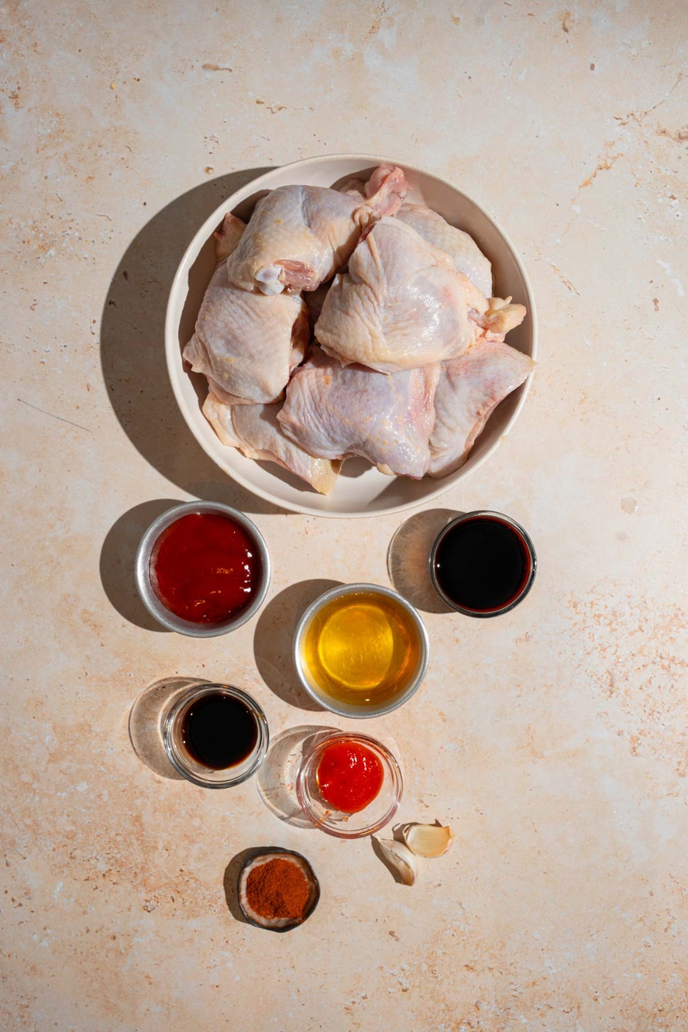 An overhead shot of several bowls in various sizes containing ingredients to make sticky chicken including uncooked chicken thighs, honey, ketchup, soy sauce, hot sauce, Worcestershire sauce, and seasonings.