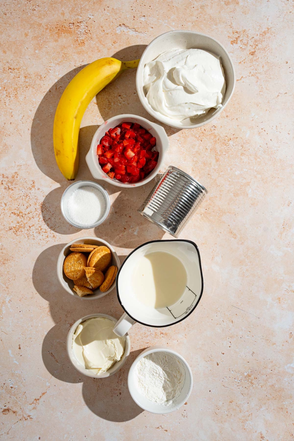An overhead shot of several bowls in various sizes containing ingredients to make strawberry banana pudding including banana, sliced strawberries, cool whip, vanilla wafer cookies, condensed milk, cream cheese, and milk.