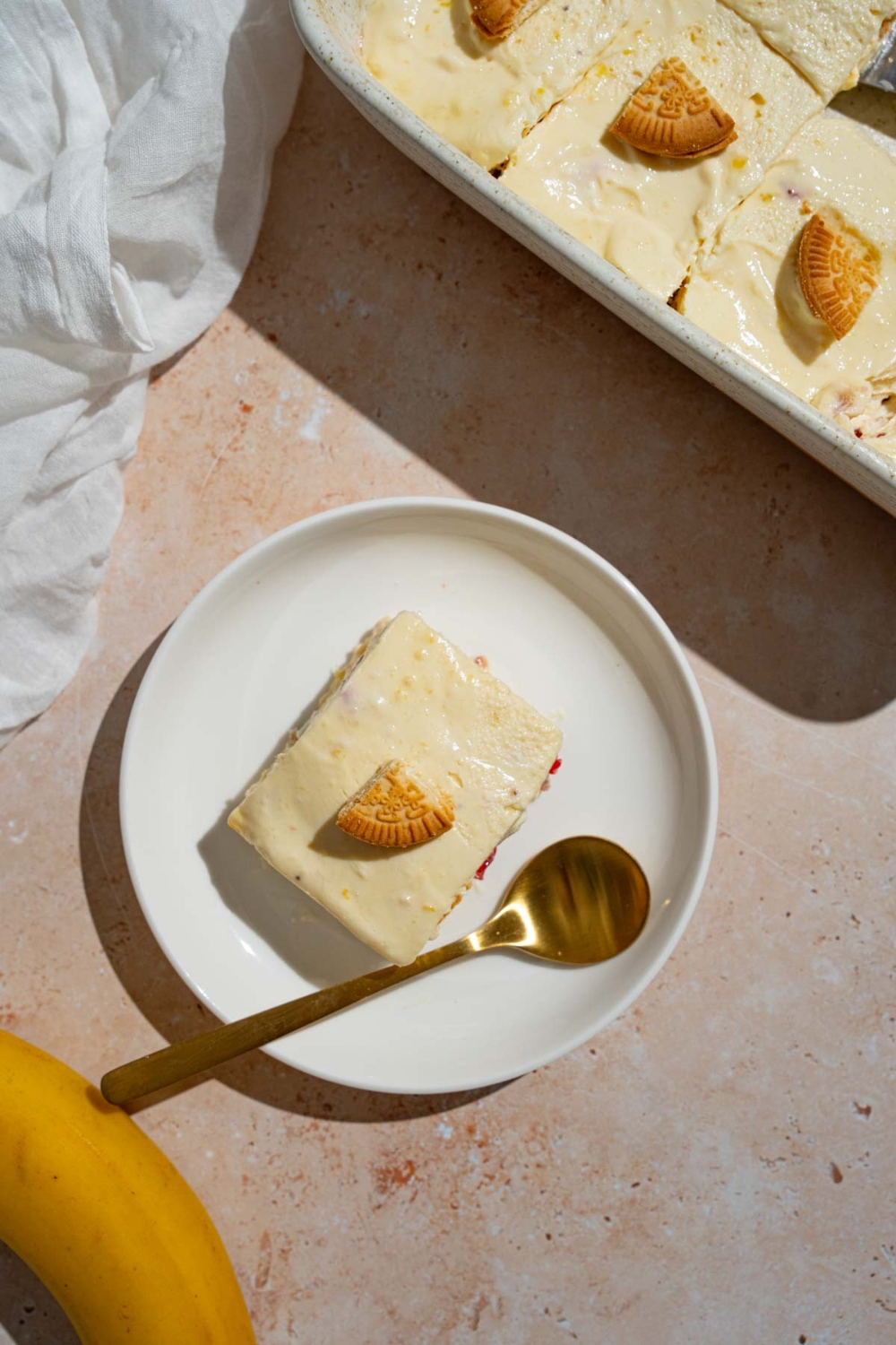 A slice of strawberry banana puding topped with a piece of a vanilla cookie. The pudding is on a white plate with a spoon. The plate is on a tan counter with a baking dish with pudding.