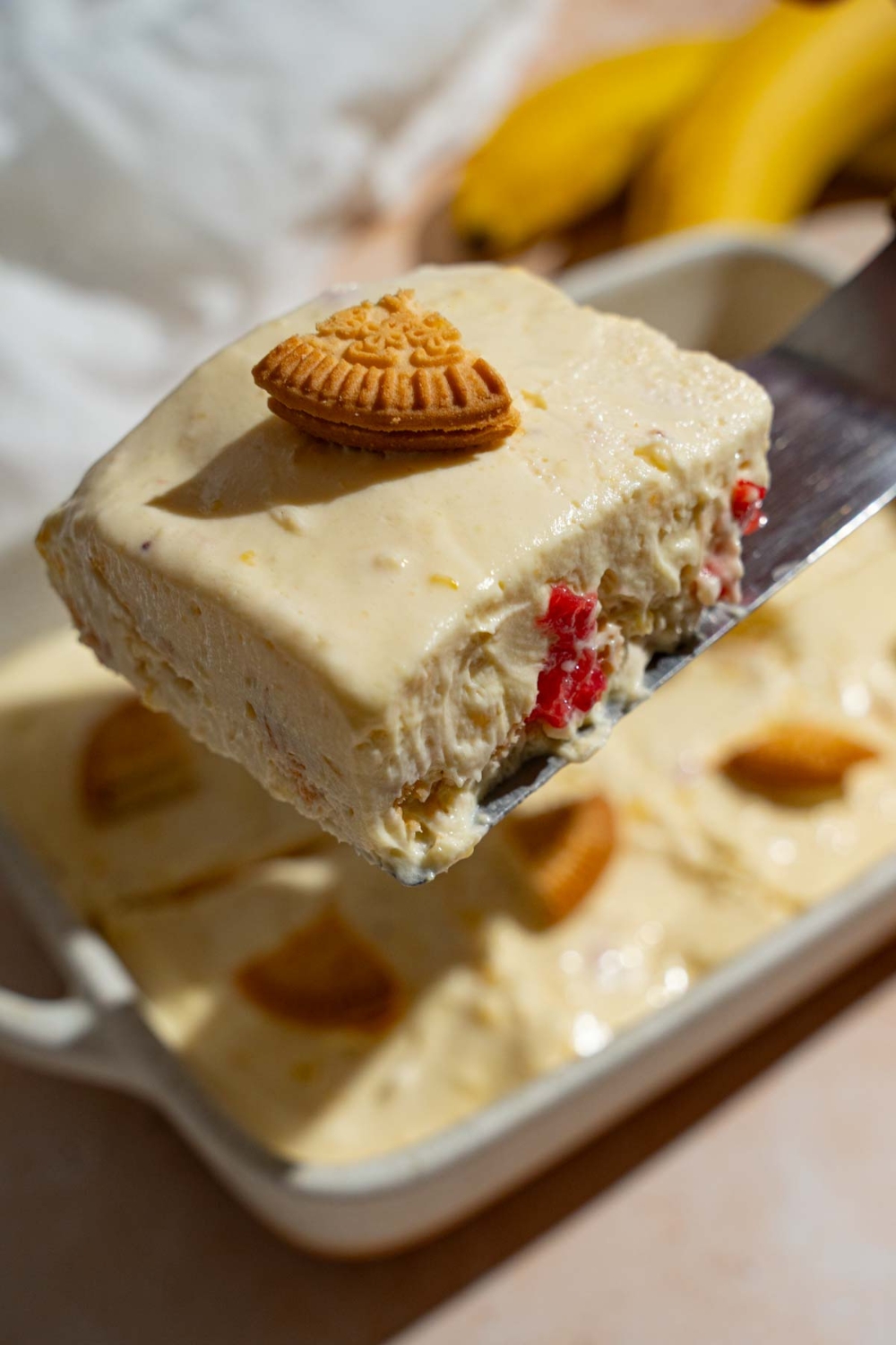A close up of a spatula holding a piece of strawberry banana pudding topped with a slice of a vanilla cookie. There is tray of pudding on a tan counter blurred in the background.