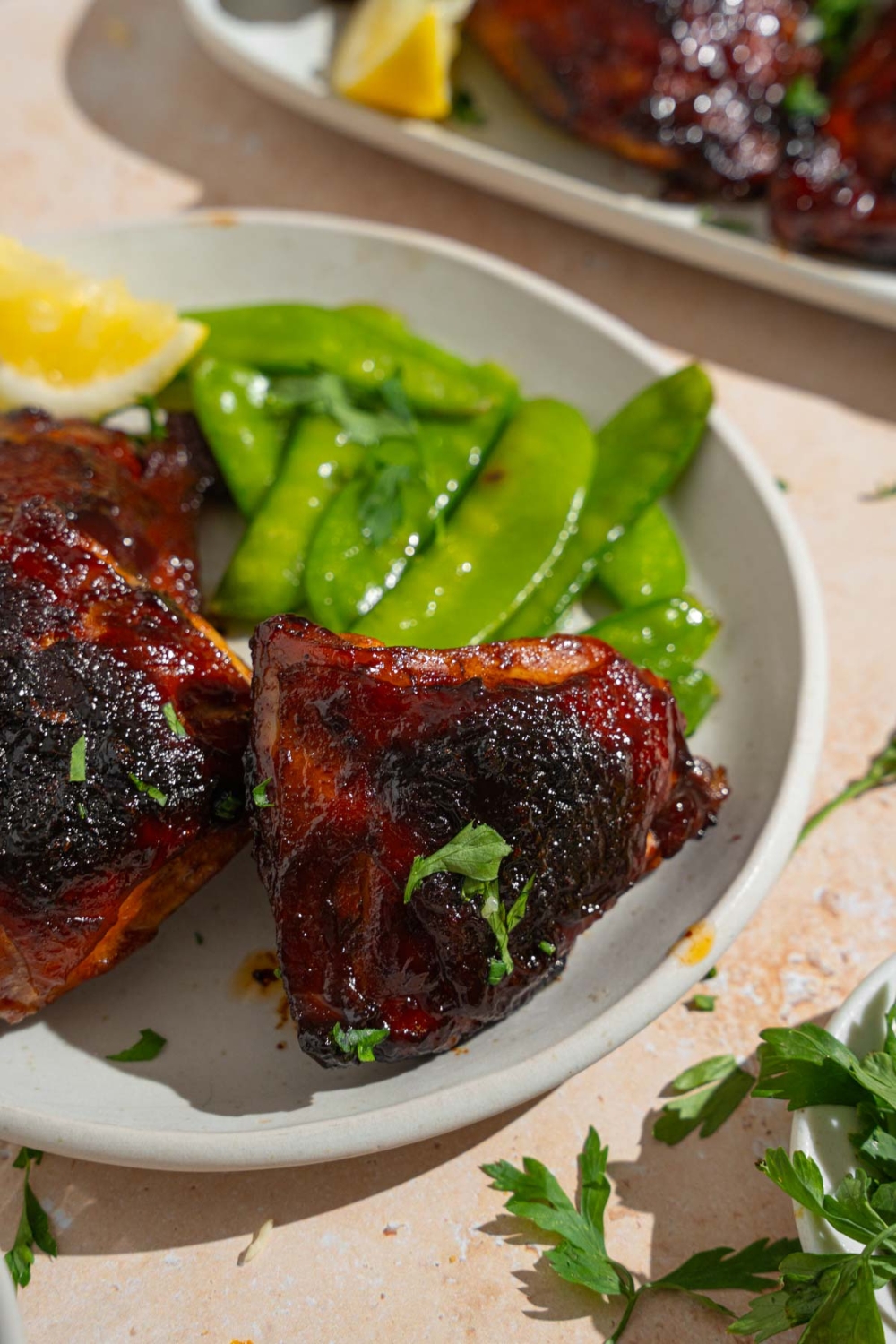 Three sticky chicken thighs served on a white plate with steamed snow peas and a lemon wedge. The plate is on a tan counter with a platter of chicken.