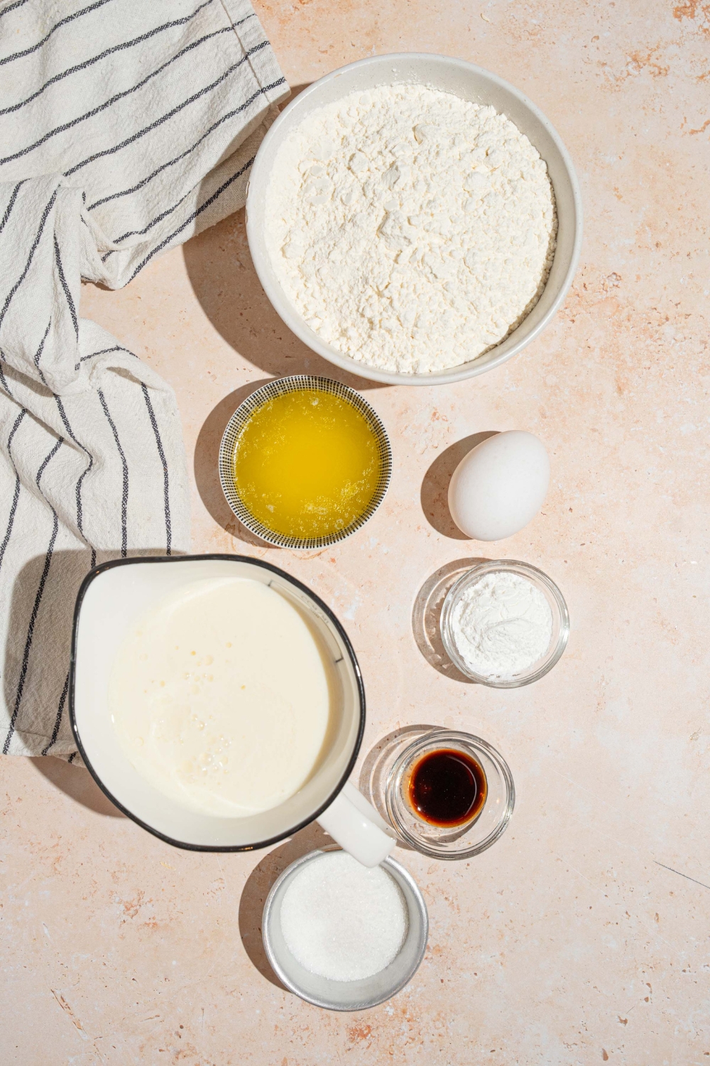 A bowl of flour, a bowl melted butter, a bowl of milk, an egg, a small bowl, baking powder, and a small old maple syrup on the white counter.