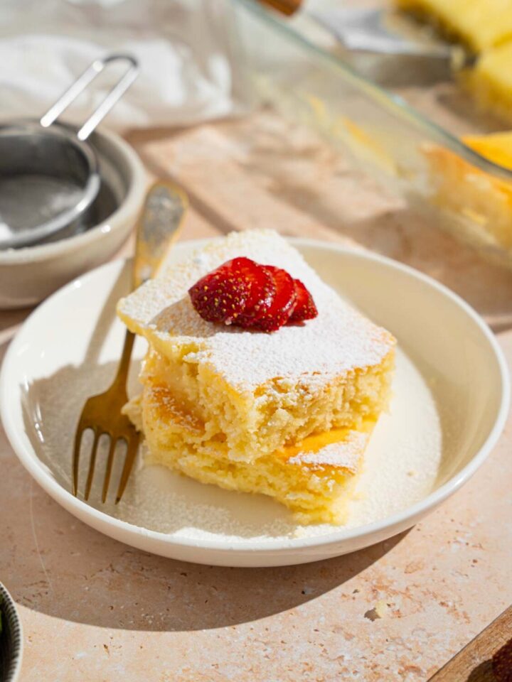A slice of neiman marcus cake on a plate with a fork on it. The cake has powdered sugar and a strawberry on top.