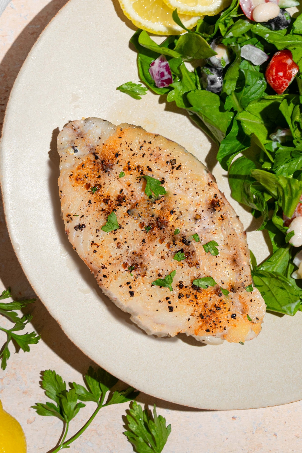 Pan seared swordfish seasoned with salt and pepper on a white plate with a side salad. The plate is on a tan counter with fresh parsley.
